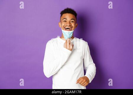 Concept of covid-19, pandemic and social distancing. Happy african-american man taking-off medical mask and breathing freely, standing over purple Stock Photo