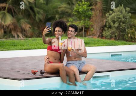 Happy multiracial young couple taking selfie in swimming pool at resort. Healthy and technology lifestyle with young people drinking fruit juice and e Stock Photo