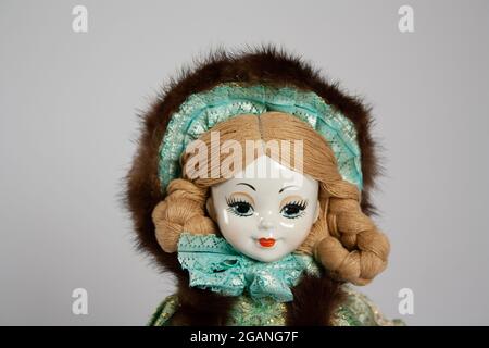 close up of the face of a porcelain doll, with braided hair and a fur cap with green lace trim on a soft gray background with copy space Stock Photo