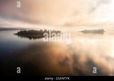 Cloudy mist sunrise over Jindabyne lake, Lion and Cub islands on calm waters - aerial snowy river. Stock Photo