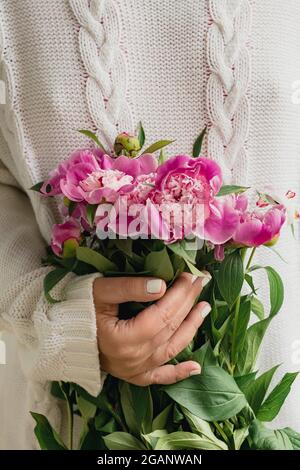 cropped young woman in white knitted sweater holding a bouquet of fresh flowers pink peonies. Stock Photo