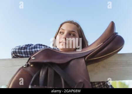 Young girl standing with her hands resting on the wooden fence in the horse ranch. Smiling and posing for the camera. Leather saddles hanging on the wooden fence in the foreground. Stock Photo