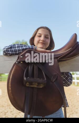 Young girl standing with her hands resting on the wooden fence in the horse ranch. Smiling and posing for the camera. Leather saddles hanging on the wooden fence in the foreground. Stock Photo