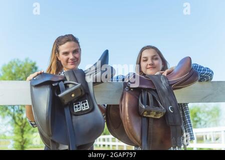 Two young girls standing with their hands resting on the wooden fence in the horse ranch. Girls smiling and posing for the camera. Leather saddles hanging on the wooden fence in the foreground. Stock Photo