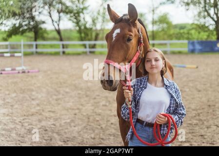 Young girl with her Bay horse standing in the stable. Girl holding horse lead rope looking at it with love. Posing for the camera. Sandy arena with wooden fence in the background. Stock Photo