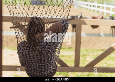 Horse owner resting on a hammock near the horse ranch during the daytime. Girl relaxing on the net swing. Horse leather saddles hanging on the wooden fence in the background. Stock Photo