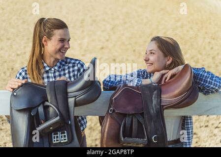 Two young girls standing with their hands resting on the wooden fence in the horse ranch. Girls in a good mood laughing and talking. Leather saddles hanging on the wooden fence in the foreground. Stock Photo
