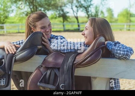 Two young girls standing with their hands resting on the wooden fence in the horse ranch. Girls in a good mood laughing and talking. Leather saddles hanging on the wooden fence in the foreground. Stock Photo
