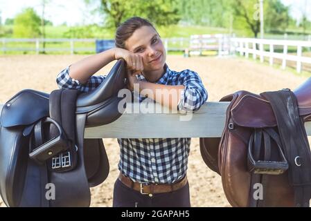 Young girl standing with her hands resting on the wooden fence in the horse ranch. Smiling and posing for the camera. Two leather saddles hanging on the wooden fence in the foreground. Stock Photo