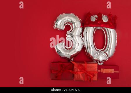 Foil silver air balloons showing number thirty placed on red background with wrapped presents for concept of birthday anniversary Stock Photo