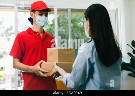 Asian delivery man wearing face mask in red uniform deliver service parcel box to woman customer at home Stock Photo