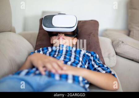 Caucasian boy lying comfortably on the sofa at home. He is using virtual reality glasses. Stock Photo