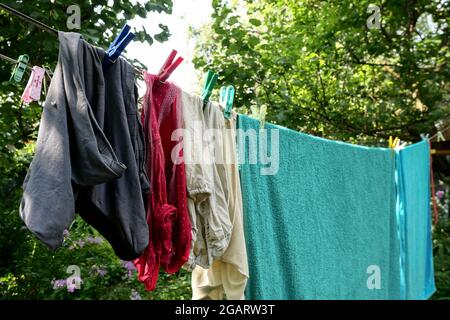 Different washed clothes is dried on rope in backyard or garden. Drying linen with multicolored clothespins. Summer sunny day. Close-up. Outdoors. Stock Photo