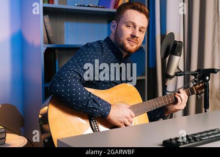 Medium shot of handsome guitarist singer man playing on acoustic guitar and singing into microphone recording song in home studio, looking at camera. Stock Photo