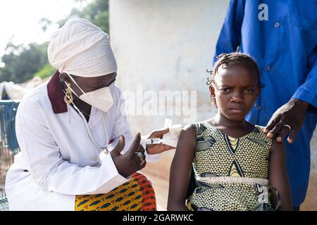 In this image, a worried little African girl is getting an influenza vaccination by a black nurse with protective face mask Stock Photo