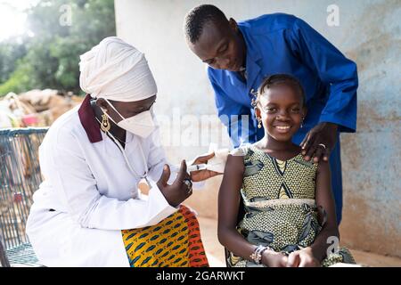 In his image, a beautiful brave little African girl with a big confident smile is getting a vaccine shot by a female doctor, with her father standing Stock Photo