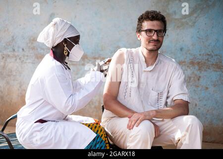 In this image, a black nurse wearing a facial mask is vaccinating a young caucasian man with a confident smile on his face against yellow fever in an Stock Photo