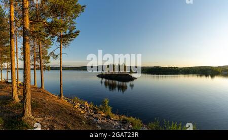 A panorama of a calm lake with small island and golden sunset evening light on the trees and forest on the lakeshore in the foreground Stock Photo