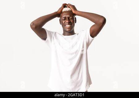 Portrait of complicated anxious african-american blond man, holding hands on head and feeling alarmed, standing over white background Stock Photo