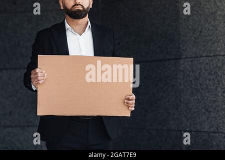 Unrecognizable male office worker in suit holding blank cardboard sign standing outdoors near business center, free space, crop. Mockup for advertisem Stock Photo