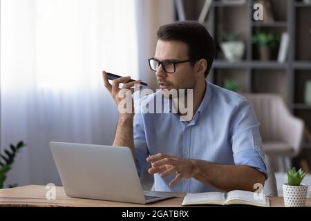 Focused male working by laptop using virtual assistant on phone Stock Photo