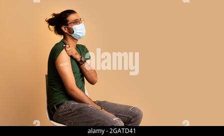 Long-haired indian man in protective face mask got vaccinated against COVID-19, showing shoulder with plaster on, sitting on chair over beige studio b Stock Photo