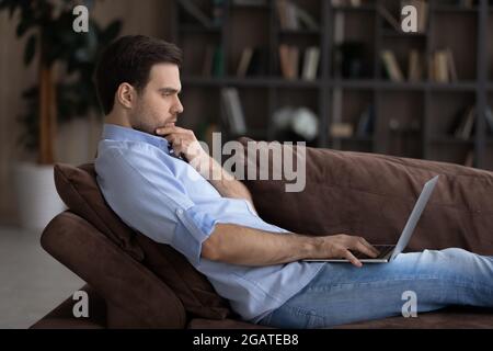 Serious young man sit on sofa engaged in computer work Stock Photo