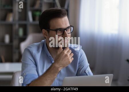 Pensive young man work at laptop online look aside think Stock Photo