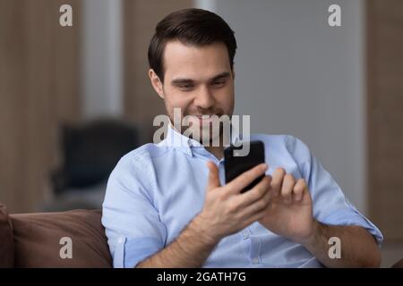 Happy young bearded guy sit on sofa enjoy using phone Stock Photo