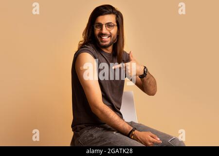 Handsome millennial long-haired indian man got vaccinated against COVID-19, pointing at shoulder with plaster on and smiling at camera, sitting on cha Stock Photo