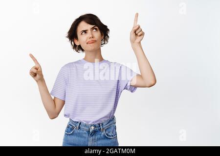 Indecisive young woman sulking, frowning serious and thoughtful, making choice, having doubts, pointing sideways but looking up, make decision Stock Photo