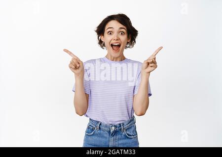 Happy girl customer, young student gasping in awe, pointing sideways, showing two banners, promo sales on two sides, smiling amazed, white background Stock Photo