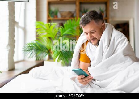 Middle aged man in bed checking his messages in smartphone after awakening. He sitting in bed under the blanket and smiling. Addiction to social netwo Stock Photo