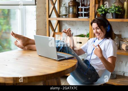 Happy smiling woman in blue headphones in front of a laptop monitor at home. She puts her feet on the table and vividly discusses the latest gossip wi Stock Photo