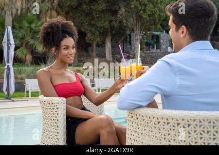 Interracial couple on vacation sitting on a table at poolside having fun holding glasses and drinking soft drinks together. Stock Photo