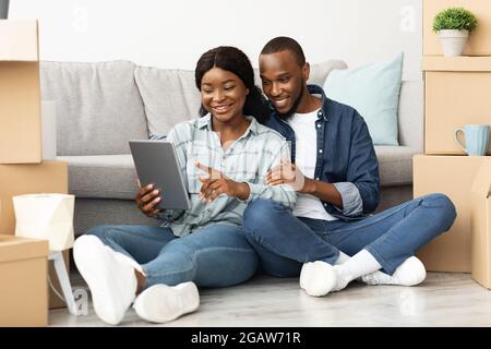 Cheerful Black Spouses With Digital Tablet Sitting Among Cardboard Boxes After Moving To New Flat, Smiling African American Couple Using Tab Computer Stock Photo
