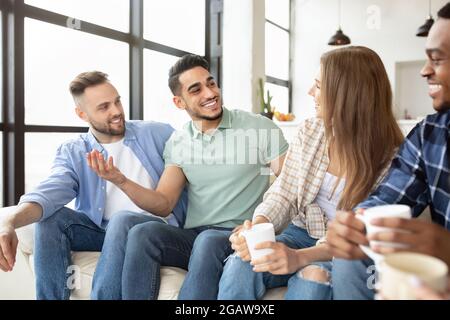 International group of friends with hot beverages chatting, sitting on sofa, having party at home. Millennial diverse students enjoying friendly conve Stock Photo