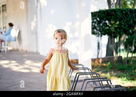 Little girl stands near a bicycle parking lot on paving stones near a green bush Stock Photo