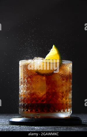 Cuba Libre or Long Island Cocktail with rum, cola and lime in glass. Soda spray on dark background. Refreshing summer drink. Stock Photo