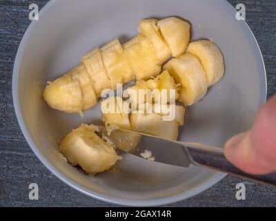 Closeup POV overhead shot of a soft ripe banana in a bowl, with the skin removed, being cut into slices with a steel knife. Stock Photo