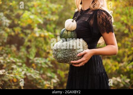 Close up of beautiful young blond woman with long straight hair, wearing black guipure dress, holding pumpkins while standing against autumn forest ba Stock Photo