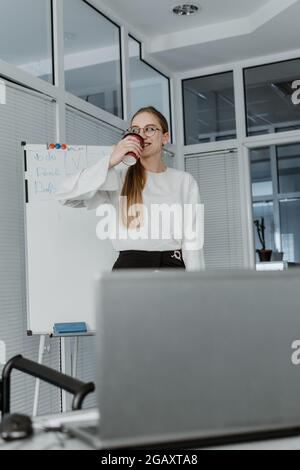 Remote Meetings, Video Conferencing. Young business woman drinking coffee and communicating with colleagues via video call on laptop in the office Stock Photo