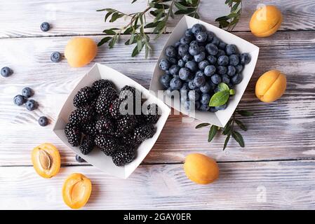 Ripe blackberries, blueberries in paper boxes and apricots on a white wooden background, summer berries, healthy food. Stock Photo