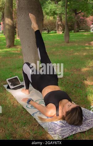 Vertical image of a young woman doing bridge yoga pose in city park in a summer sunny day. Pilates Workout Outdoor 2021. Stock Photo