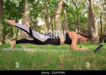 Vertical image of a young girl doing a plank pilates pose in city park in a summer sunny day. Pilates Outdoor 2021. Stock Photo