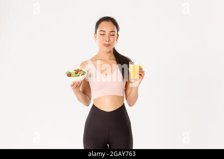 Sport, wellbeing and active lifestyle concept. Smiling cute fitness asian girl in activewear, licking lips satisfied with delicious healthy salad and Stock Photo