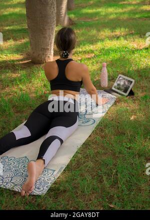 Vertical image of a young girl doing the cobra pose while she is relaxing outdoors in city park in a summer sunny day. Pilates 2021 Stock Photo