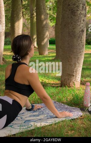 Vertical image of a young girl doing the cobra pose while she is relaxing outdoors in a summer sunny day. Pilates Outdoor 2021. Stock Photo