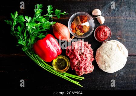 Raw Turkish Lahmacun Ingredients on a Dark Wood Background: Raw ground beef, vegetables, spices, and dough on a wooden table Stock Photo