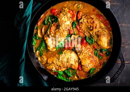 Italian Skillet Chicken with Tomatoes and Mushrooms in a Cast Iron Skillet: Chicken cutlets with spinach, mushrooms, and tomatoes in a dark wood table Stock Photo